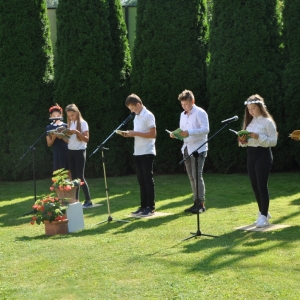 Sześcioro uczniów stojących przed mikrofonami czytają fragmenty Balladyny. Uczniowie stoją na trawniku a za nimi znajdują się zielone drzewa. Na przodzie dwie donice z czerwonymi kwiatami.