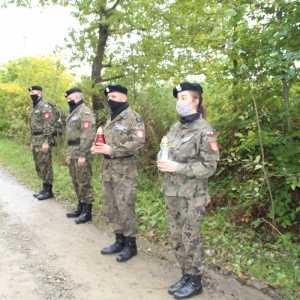 Na leśnej drodze stoją ubrani w mundury trzech żołnierzy i żołnierka Jednostki Strzeleckiej 2222. Dwoje z nich trzyma zapalone znicze. W tle zieleń i drzewa.